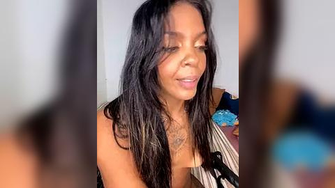 Media: Video of a Black woman with long, straight black hair, medium skin tone, and a tattoo on her chest. She is wearing a sleeveless top with a pattern, smiling softly. Background is blurred.