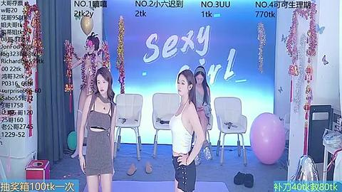 Media: Video of two Asian women in revealing outfits on a stage with \"Sexy Girl\" sign, surrounded by balloons and audience chairs, capturing a lively and vibrant atmosphere.