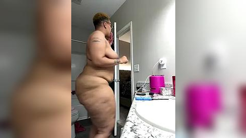 Media: Video of a nude, plus-size woman with short blonde hair and glasses, leaning against a bathroom counter with pink and blue toiletries.