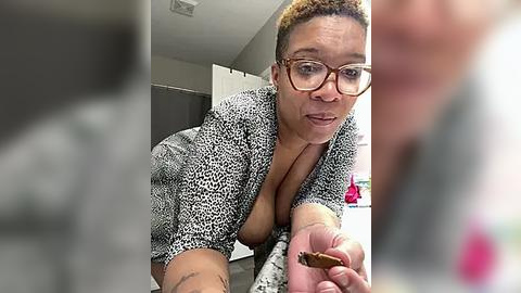 Media: Video of a middle-aged Black woman with short, curly hair, glasses, and a leopard print robe, eating a sandwich. Background shows a bathroom with white walls and a pink bottle on the counter.