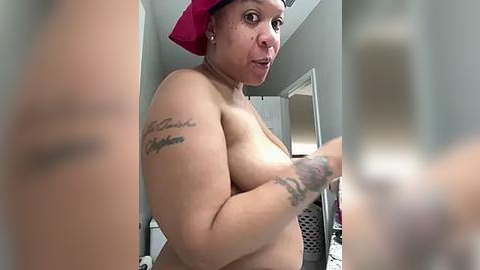 Media: A video of a topless, dark-skinned woman with a medium build, wearing a pink headscarf and holding a phone. She has tattoos on her arm and is in a bathroom.