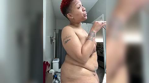 Media: Video of a nude, plus-sized, African American woman with short red hair, tattoos, and a curvy physique, standing in a bathroom, holding a phone.