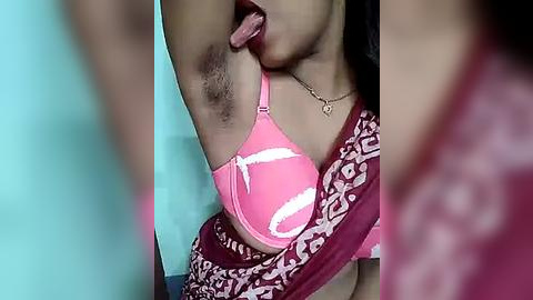 Media: Video of a woman with light brown skin, wearing a pink bra with white designs, licking her armpit, her tongue visible, with a blurred background.