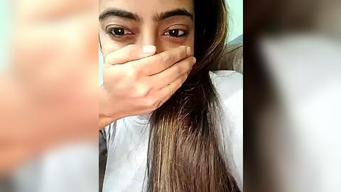Media: Video of a young woman with long, dark hair covering her mouth, wearing a white shirt, taken indoors with blurred background.