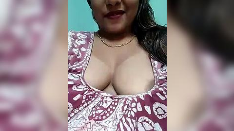 Media: Video of a woman with medium skin tone, dark hair, and large breasts, wearing a low-cut, floral-patterned blouse, standing against a teal background.
