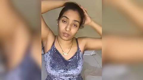 Media: Video of a young woman with medium brown skin and long dark hair, wearing a sleeveless blue patterned top, standing in a bedroom with a white bed. Her hands are on her head.