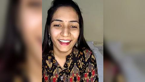 Media: Video of a smiling South Asian woman with medium brown skin, straight black hair, and traditional red bindi. She wears a colorful, patterned blouse with silver earrings. The background is blurry, suggesting an indoor setting.