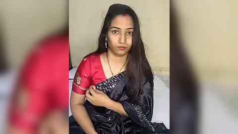 Media: Video of a young South Asian woman in a red sari with gold embroidery, seated on a bed in a beige room. She has long black hair and a neutral expression.