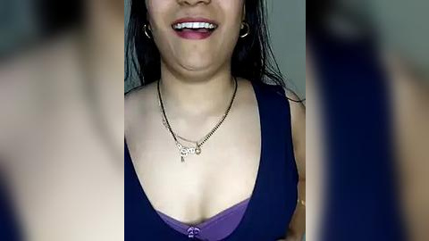 Media: Video of an Asian woman with medium skin tone, wearing a purple bra, a black tank top, and a silver necklace. She has long black hair and is smiling.