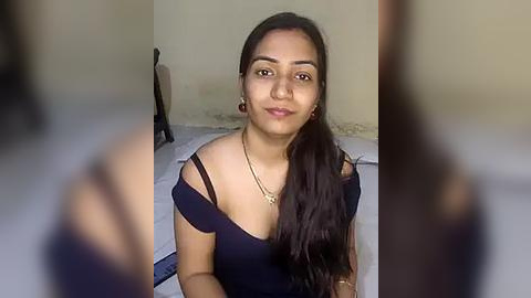 Media: A video of a young woman with medium brown skin, long dark hair, and a slender build, wearing a black off-shoulder top, seated indoors against a plain beige wall.