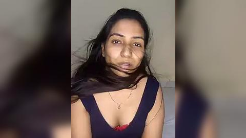 Media: Video of a South Asian woman with medium-brown skin and long black hair, wearing a deep V-neck black dress with a red bra peeking out, and a gold necklace.