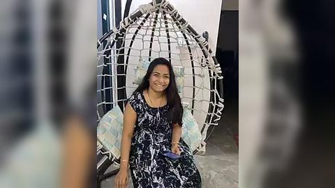 Media: Video of a smiling South Asian woman with long black hair, wearing a black and white patterned dress, sitting in a white woven hanging chair. Background shows a blurry room with concrete floor and a doorway.