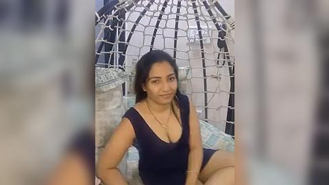 Media: Video of a smiling, dark-skinned woman with long dark hair, wearing a black V-neck dress, sitting in a white wicker swing chair against a blue wall, indoors.