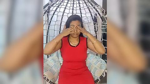 Media: Video of a woman with medium brown skin, medium build, and medium-length black hair, wearing a red dress, covering her eyes with her hands, seated in a white hammock chair.