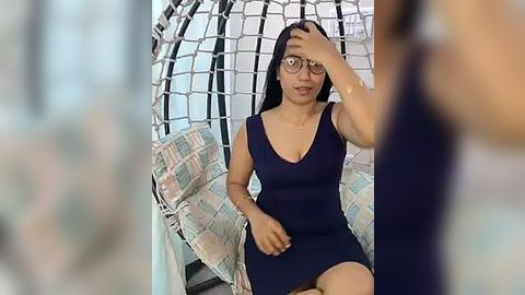 Media: A video of a woman with long black hair, wearing glasses, a dark blue dress, and seated in a hanging egg chair, with blurred background.