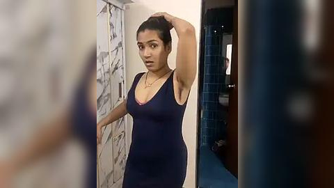 Media: A video of a woman with medium brown skin and dark hair in a tight navy dress, taken from a bathroom mirror. The background shows a tiled wall and a blue carpet.