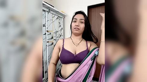 Media: A video of a curvy woman with medium skin tone, long black hair, and wearing a purple bra and pink sari, standing indoors.