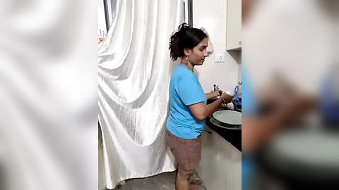 Media: Video of a South Asian woman with medium brown skin and black hair tied in a ponytail, wearing a blue shirt and brown shorts, washing dishes in a small kitchen with white curtains and a white sink.