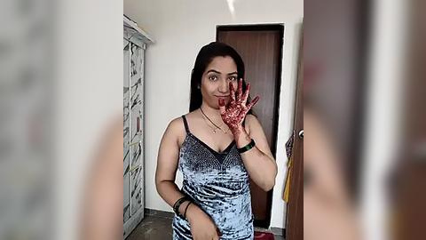 Media: Video of a woman with long black hair, wearing a blue tie-dye dress, standing in a doorway with a bloody handprint on her face, looking at the camera.