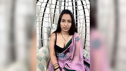 Media: Video of a young Asian woman with medium skin tone, long black hair, wearing a pink sari with black lace bra, seated in a white and black woven chair.