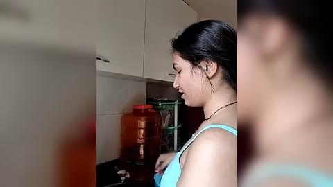 Media: Video of a woman with long black hair, wearing a blue tank top, filling a large orange water jug in a modern kitchen with white cabinets and a green metal rack.