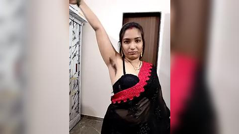 Media: Video of a South Asian woman with medium brown skin and dark hair, wearing a black sari with red trim, standing in a narrow, white-walled hallway with a wooden door behind her.