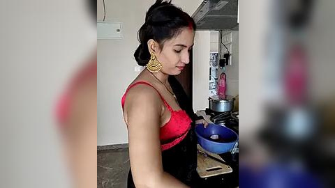 Media: Video of a young woman with medium brown skin, wearing a red lace bra, gold hoop earrings, and a black sari, cooking in a cluttered kitchen with a blue bowl on the counter.