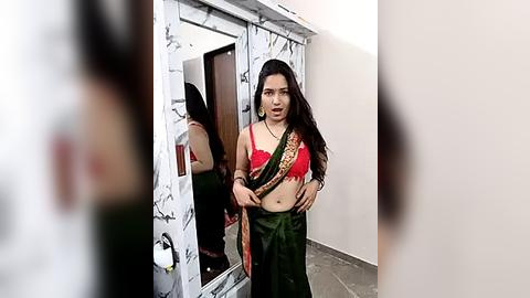 Media: A video of a South Asian woman with long black hair, wearing a red bra and green sari, standing in front of a mirror. The room has white marble floors and a wooden door.