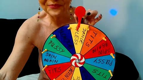 Media: A video of a topless woman with fair skin and blonde hair, holding a colorful, candy-themed pinwheel spinner with naughty words like \"Spin Again\" and \"Pussy.\" The background is a plain wall.