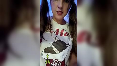 Media: Video of a young woman with long brown pigtails, wearing a white \"Maz\" t-shirt featuring a cat graphic, in a dimly-lit room with blurred background.