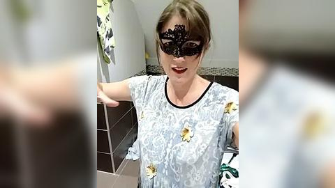 Media: Video of a woman with a black lace mask, light brown hair, and a white floral blouse, smiling in a modern bathroom with tiled walls.