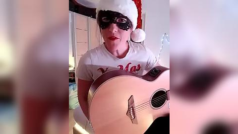 Media: Video of a man with pale skin, wearing a white T-shirt, black mask, and Santa hat, playing an acoustic guitar in a dimly lit room.