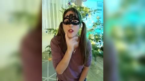 Media: Video of a young woman with fair skin, long brown hair in pigtails, wearing a black lace mask and purple top, holding her chin, in a garden with greenery and a blue sky.