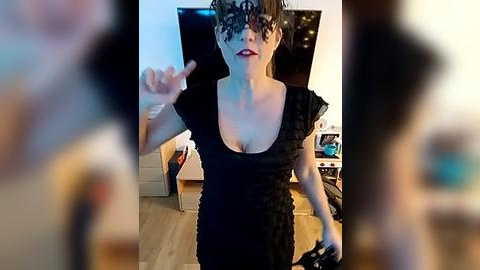 Media: A video of a woman in a black lace mask, red lipstick, and a black dress, gesturing with her hand, indoors with a blurred background featuring a TV, furniture, and a lamp.