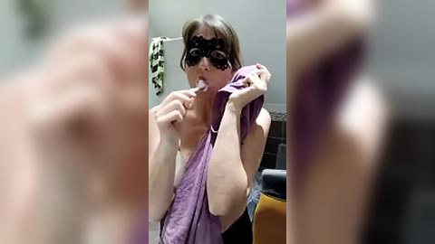 Media: Video of a woman in a purple dress, wearing a black mask and brushing her teeth in a bathroom with a towel and mirror.