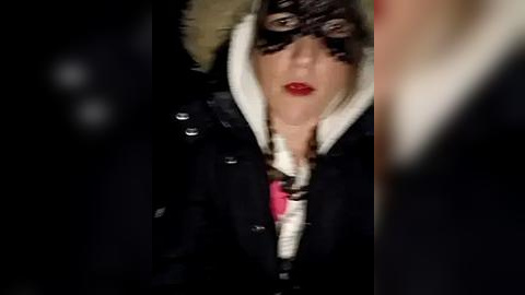 Media: Video of a person with a pale complexion, wearing a black lace mask, a black coat with white fur trim, and red lipstick, set against a dark background, creating a dramatic, mysterious mood.