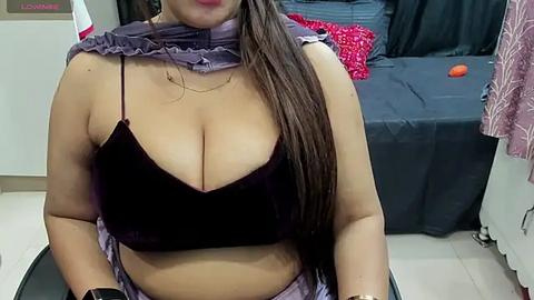 Media: A video of a plus-sized woman with long brown hair, wearing a black velvet bralette, purple scarf, and patterned skirt, sitting in a bedroom with a dark bed and pink pillow.