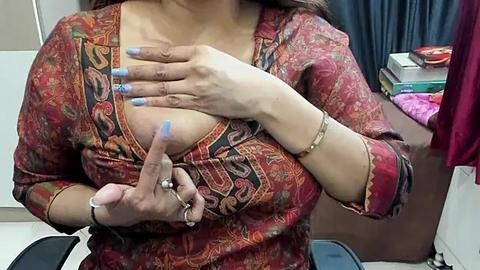 Media: A video of a woman with light brown skin, wearing a vibrant red and orange patterned blouse, exposing her left breast. She has blue-painted nails and a gold bracelet on her wrist. The background shows a cluttered room with a bed and various items.