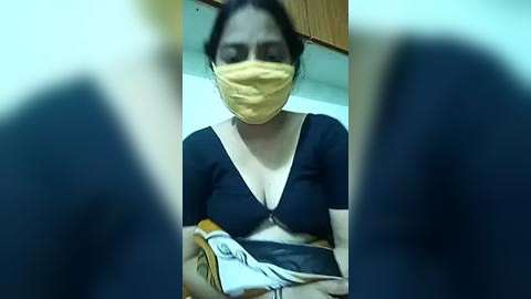 Media: Video of a woman with a yellow face mask, dark hair, and a black-and-white outfit, holding a yellow and white book, taken from a low angle, with blurred foreground.