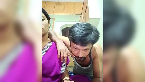 Media: Video of a man with short black hair in a gray tank top kissing a woman in a purple sari. Background shows a wooden closet and beige walls.