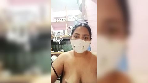 Media: Video of a woman with medium skin tone, dark hair, and a white surgical mask, seated in a kitchen with pastel walls and visible utensils, topless, revealing medium-sized breasts.