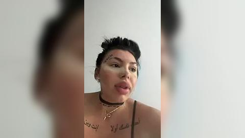 Media: Video of a woman with dark hair in a messy bun, wearing a black choker and tattoos on her chest, licking her lips. Background is blurred.