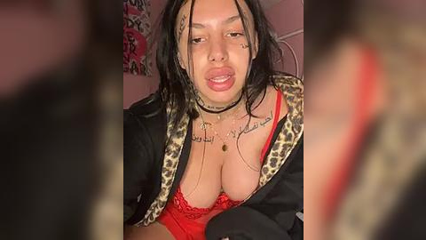 Media: Video of a young woman with medium skin tone and long black hair, wearing red lace lingerie and a leopard-print jacket, with tattoos and piercings, indoors.