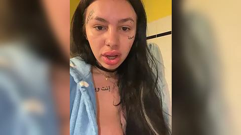 Media: A video of a young woman with long black hair, wearing a light blue robe, and multiple tattoos on her face and neck, taken in a bathroom.