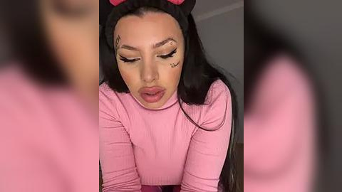 Media: A video of a young woman with long black hair, wearing a pink ribbed turtleneck and a black headband with pink cat ears, leaning forward with closed eyes, and small heart-shaped tattoo above her right eye.