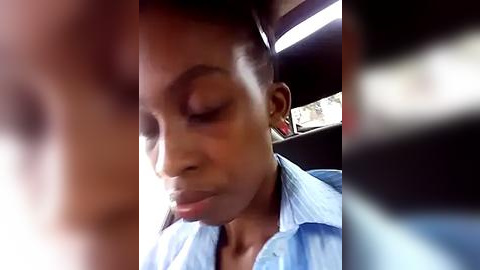 Media: Video of a young Black woman with short hair, wearing a light blue shirt, sitting in a car with blurry background. She has a serene expression, possibly sleeping or deep in thought.