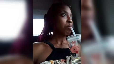 Media: Video of a young Black woman with medium brown skin and red hair, wearing a black floral-patterned top, taking a selfie while drinking from a clear plastic cup with a straw.