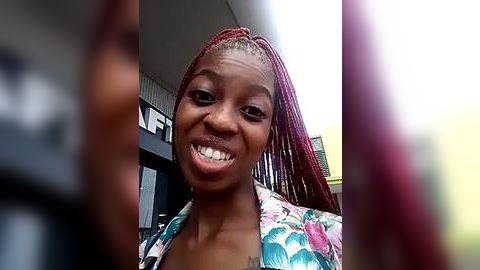 Media: Video of a young Black woman with dark skin, smiling widely, wearing red braided hair and a floral blouse. She stands in a modern indoor setting with blurred background details.