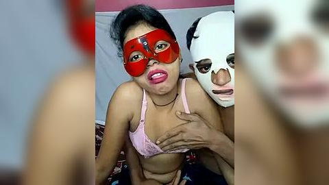 Media: Video of a young woman with dark hair, wearing a red mask and pink bra, being held by a man with a white mask, in a room with red and white walls.