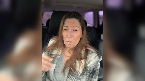 Media: Video of a middle-aged woman with long, straight brown hair, wearing a plaid shirt, smoking a cigarette in a car. The background shows a blurred, dark interior with seats.